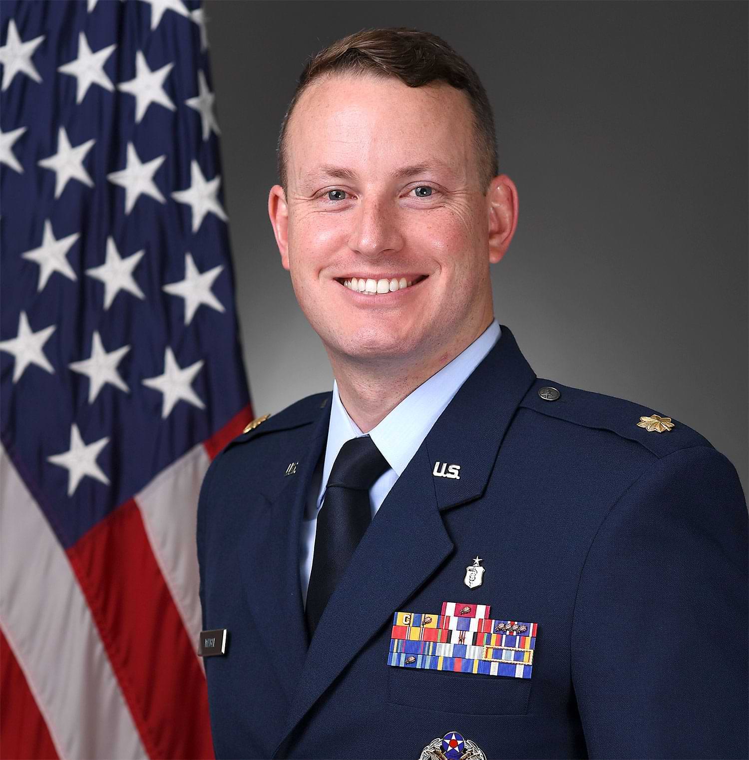 Square portrait photograph of Timothy Weigle, PharmD smiling in his United States Air Force outfit while posing in front of the American flag