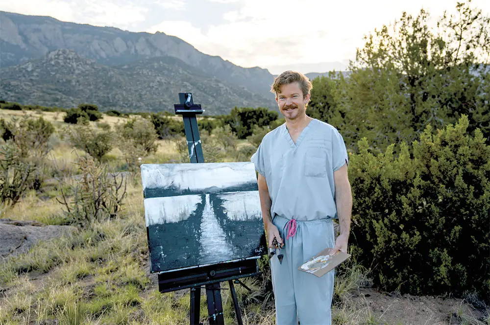 Artist standing outside dressed in scrubs next to easel with painting on it