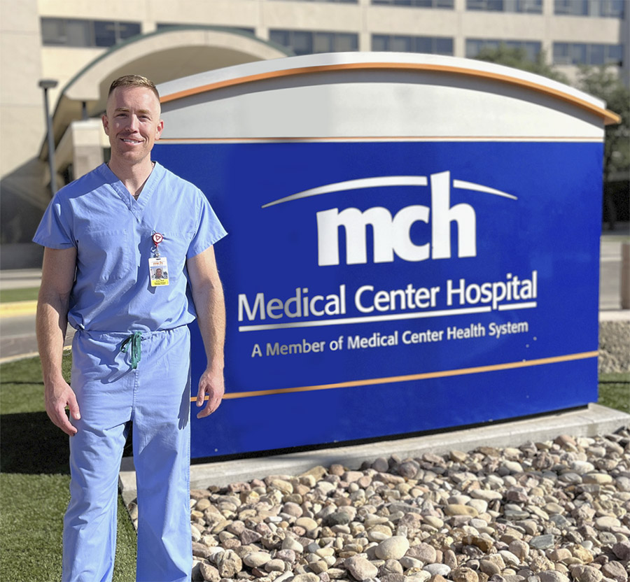 A portrait photograph of Nathanael Longacre, PA-C, (Health Professions ’21) smiling in his blue medical gown outfit posing outside in front of the blue Medical Center Hospital sign in Odessa, Texas