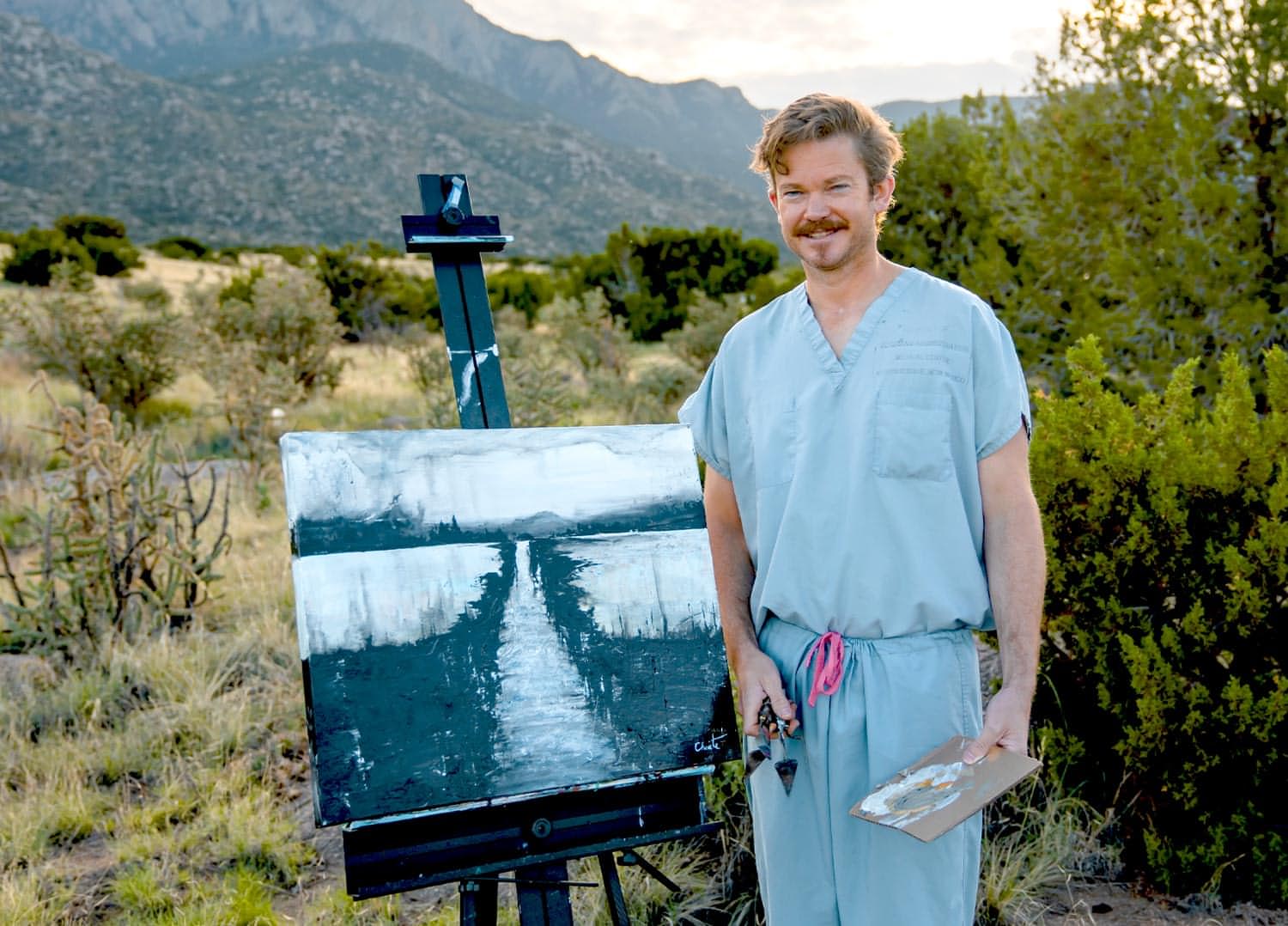 Bevan Choate wearing medical scrubs and standing among hills and shrubs, smiles next to a palette knife painting