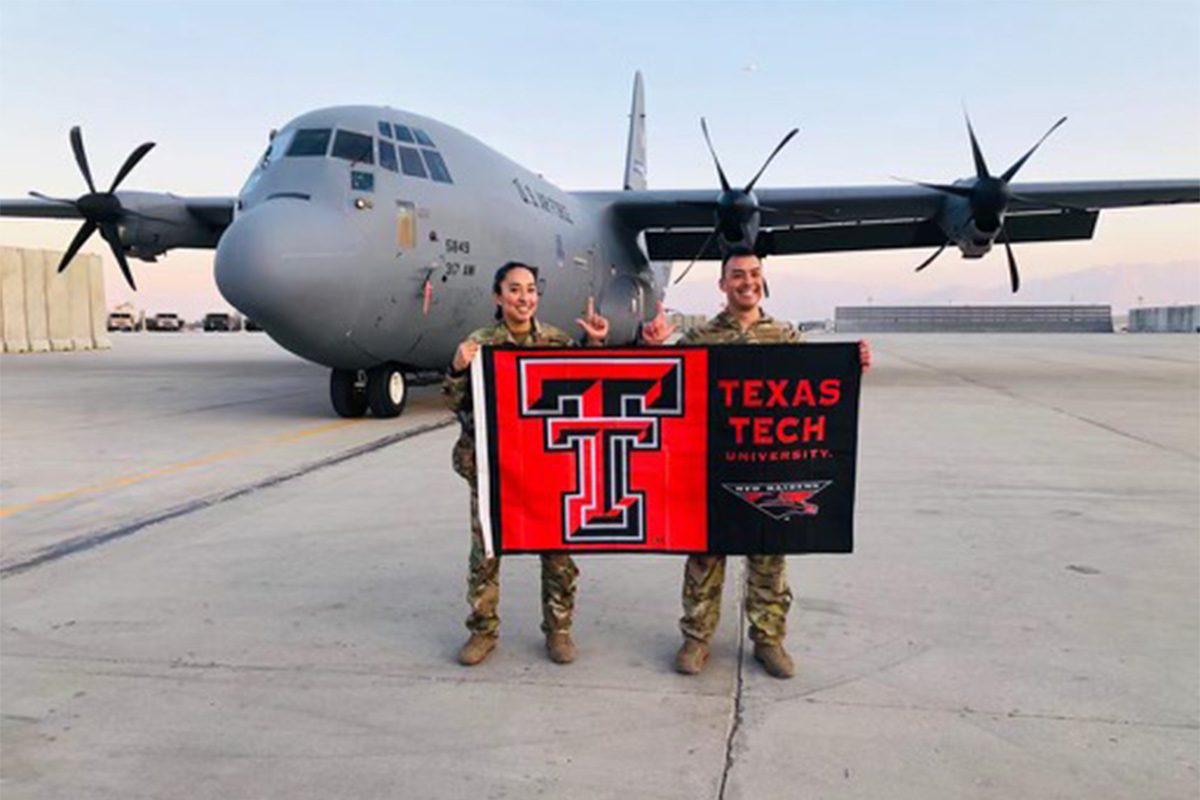 2 soldiers holding school flag in front of military cargo plane