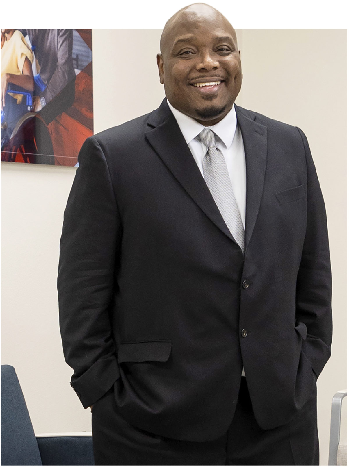 A portrait photograph of Chris Townsend (PhD, LPC, AADC – Your Life Behavorial Health and Wellness Clinic director) smiling in a black suit and white button-up dress shirt underneath with light grey tie as he poses with his arms placed inside the front pockets of his black dress pants