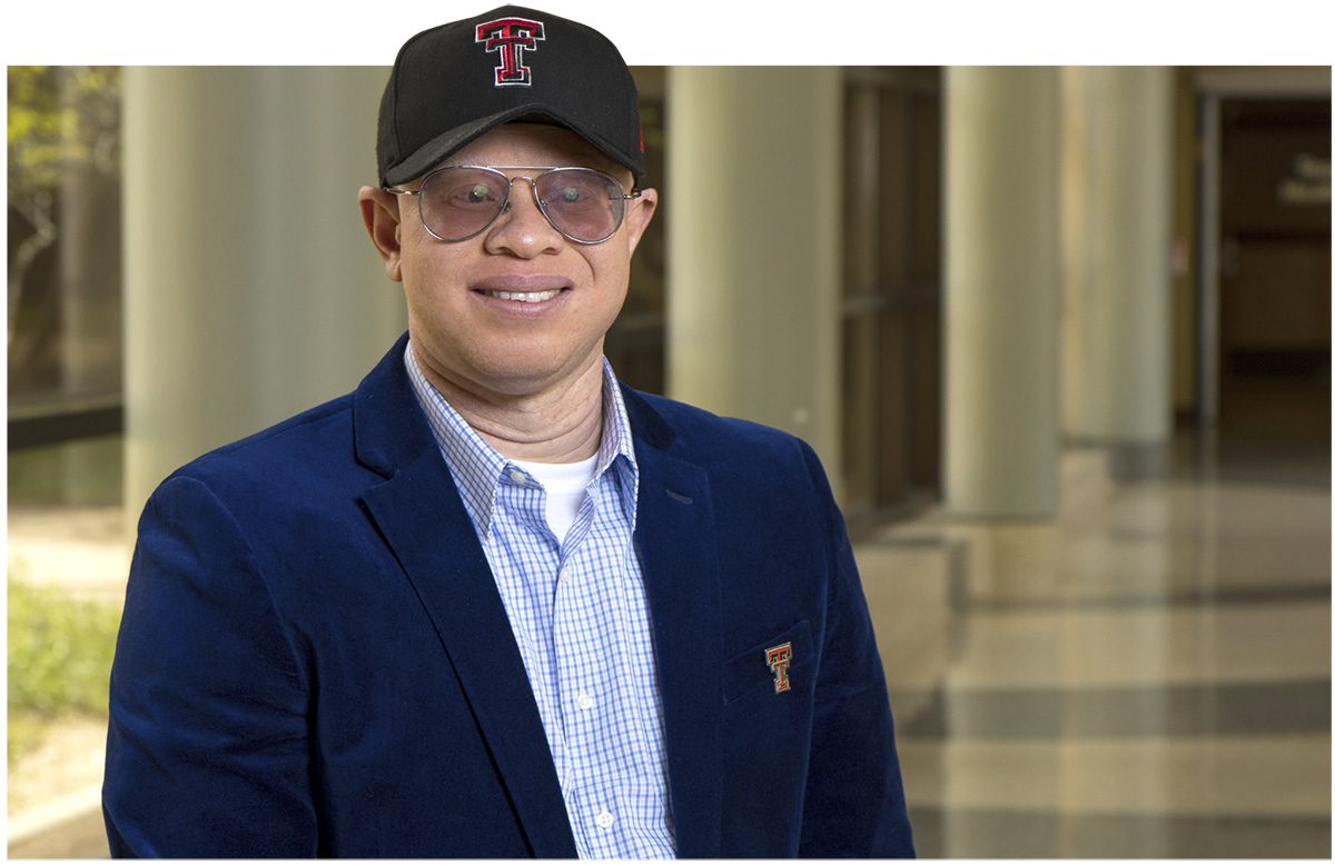 A landscape photograph of Duke Appiah (PhD, MPH – associate professor for the Julia Jones Matthews School of Population and Public Health) smiling in a dark blue suit with the Texas Tech University logo stitched on top of a pocket and light baby blue tile-shaped line pattern style button-up dress shirt underneath wearing see through tinted glasses and a black and red Texas Tech University hat