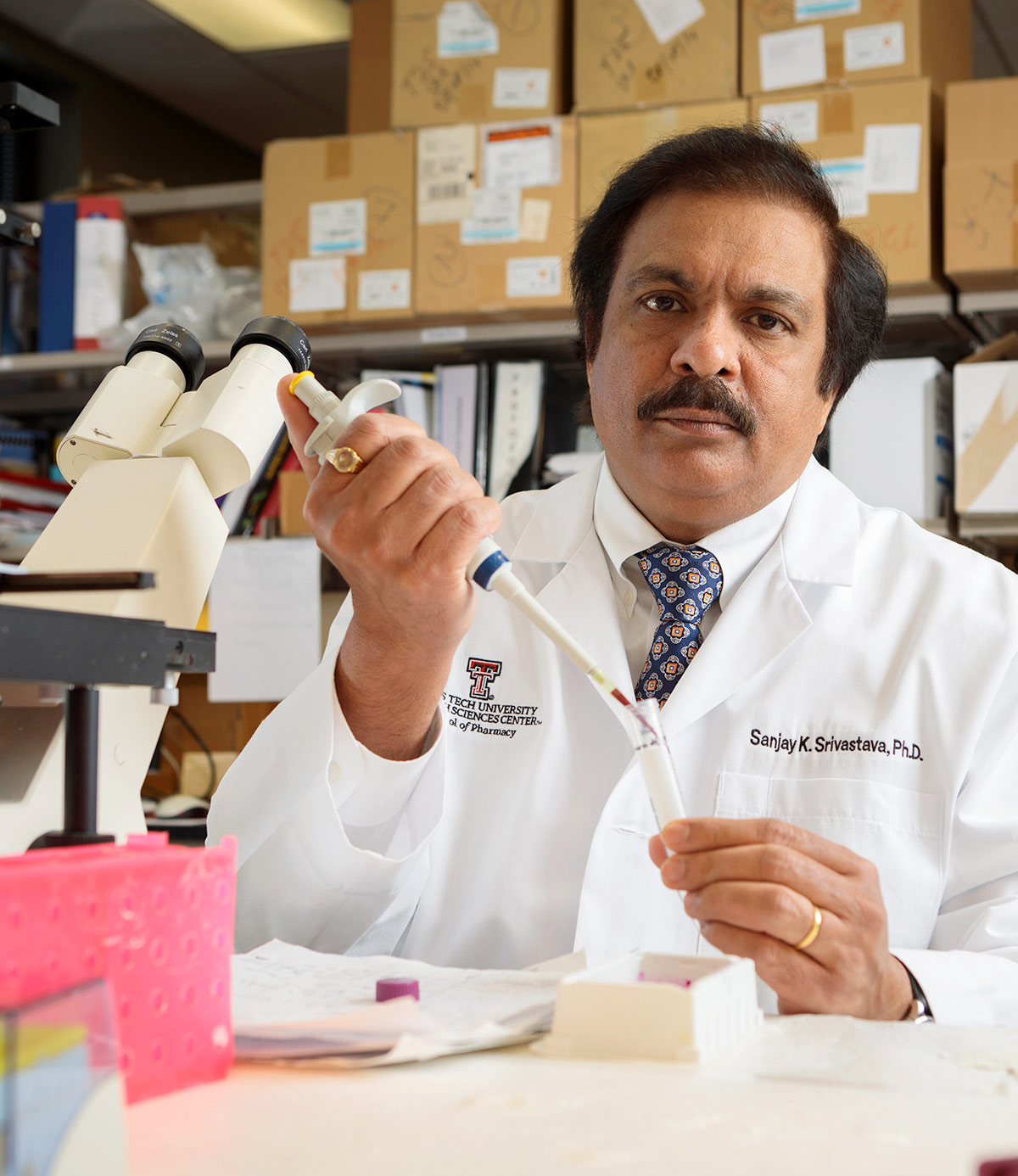 A portrait photograph of Sanjay Srivastava, PhD wearing a lab coat while using a pipette at a desk