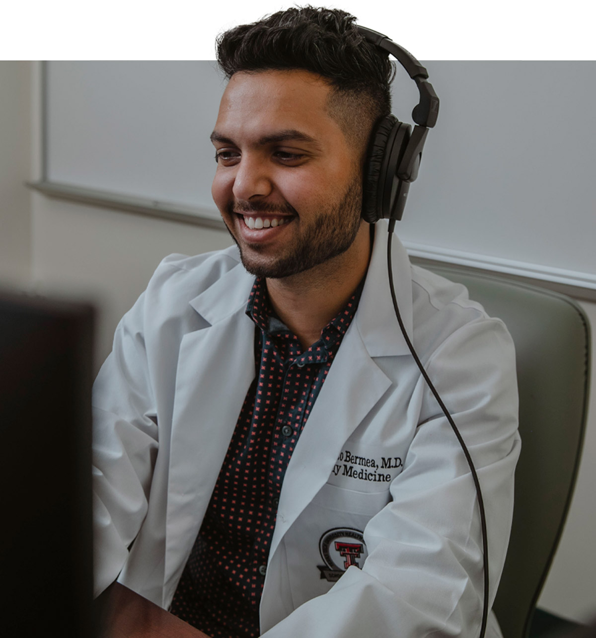 a man in a Texas Tech lab coat sits at a computer, smiling and wearing headphones