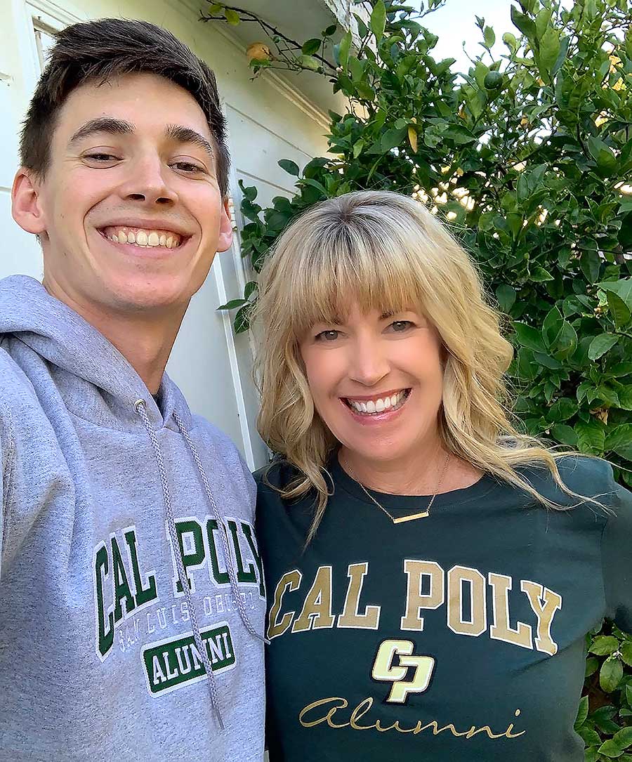 Portrait photograph perspective of Traci Henegar (in a dark forest/emerald green colored Cal Poly San Luis Obispo Alumni sweatshirt and gold colored necklace) and her son, Davis Henegar (in a grey colored Cal Poly San Luis Obispo Alumni sweater) smiling and posing for a picture together outside