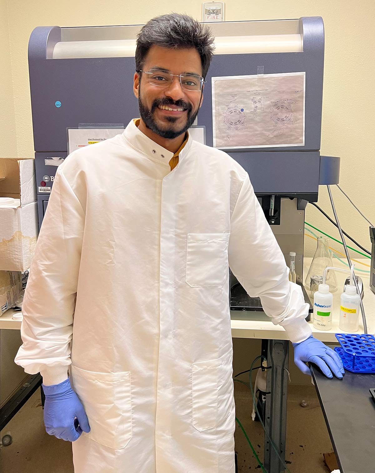 Portrait photograph perspective of Shreyas Gaikwad (a PhD candidate in pharmaceutical sciences) smiling in a white laboratory gown with blue surgical gloves as he poses for a picture by leaning his left hand on a black table with other scientific equipment nearby