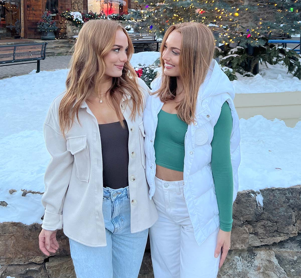 Portrait photograph perspective of Abigail Rickli and her twin sister, Avery Rickli outside in a snowy cold Christmas village location looking at each other in the eyes as they both smile as one of the sisters is wearing a black long-sleeve shirt with an open tan colored cardigan button-up shirt, blue denim jeans, and a pearl colored necklace and the other sister is wearing a white puffy jacket with a green long sleeve shirt plus white jeans