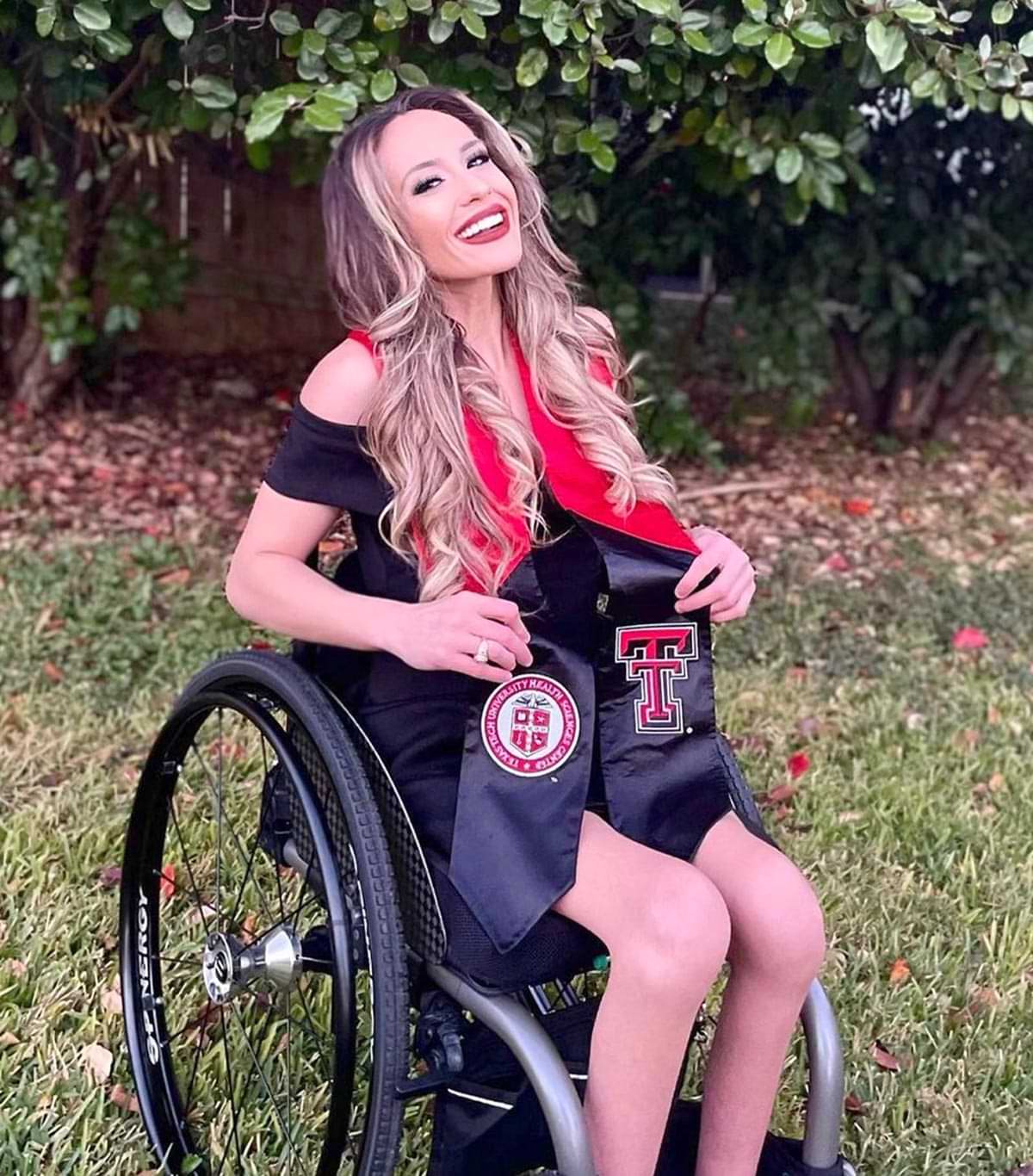 Meagan Prehn, BSN, RN photographed in her wheelchair outside near shrubs, she wears a black dress and a Texas Tech grad stole beneath her long curly hair, she smiles at the camera