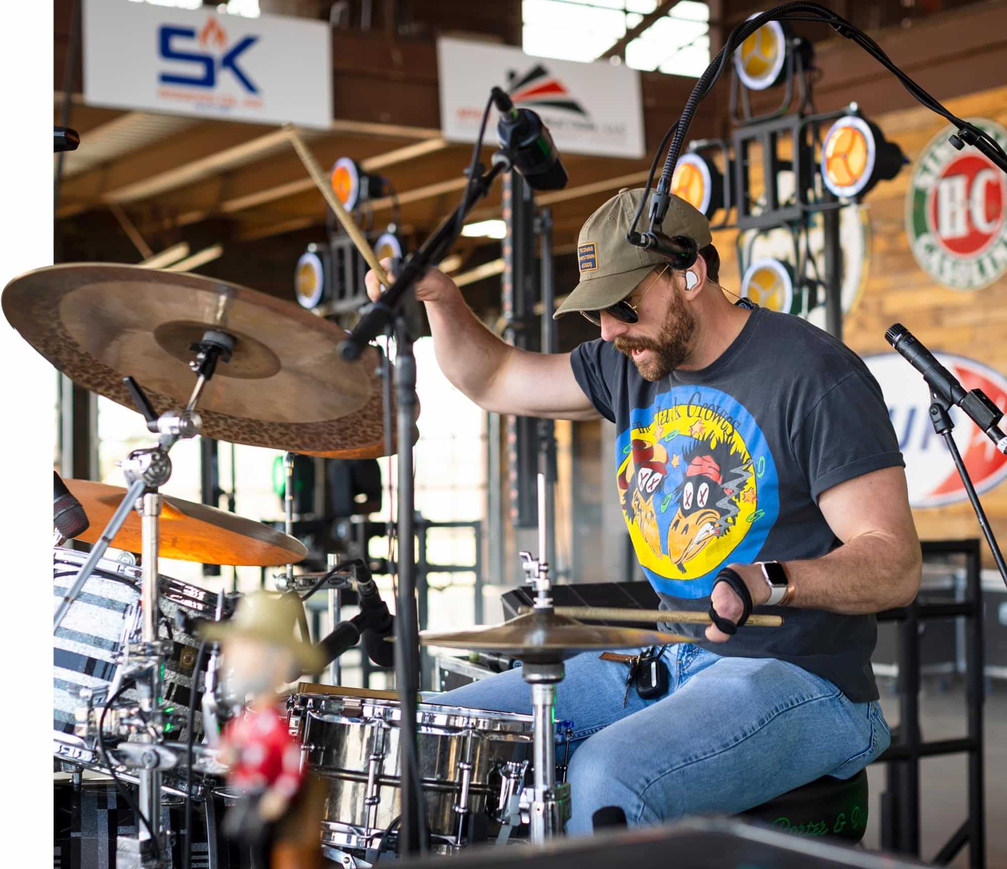 close up of Jason Albers performing drums on a stage wearing sunglasses, a green cap, a graphic t-shirt and jeans