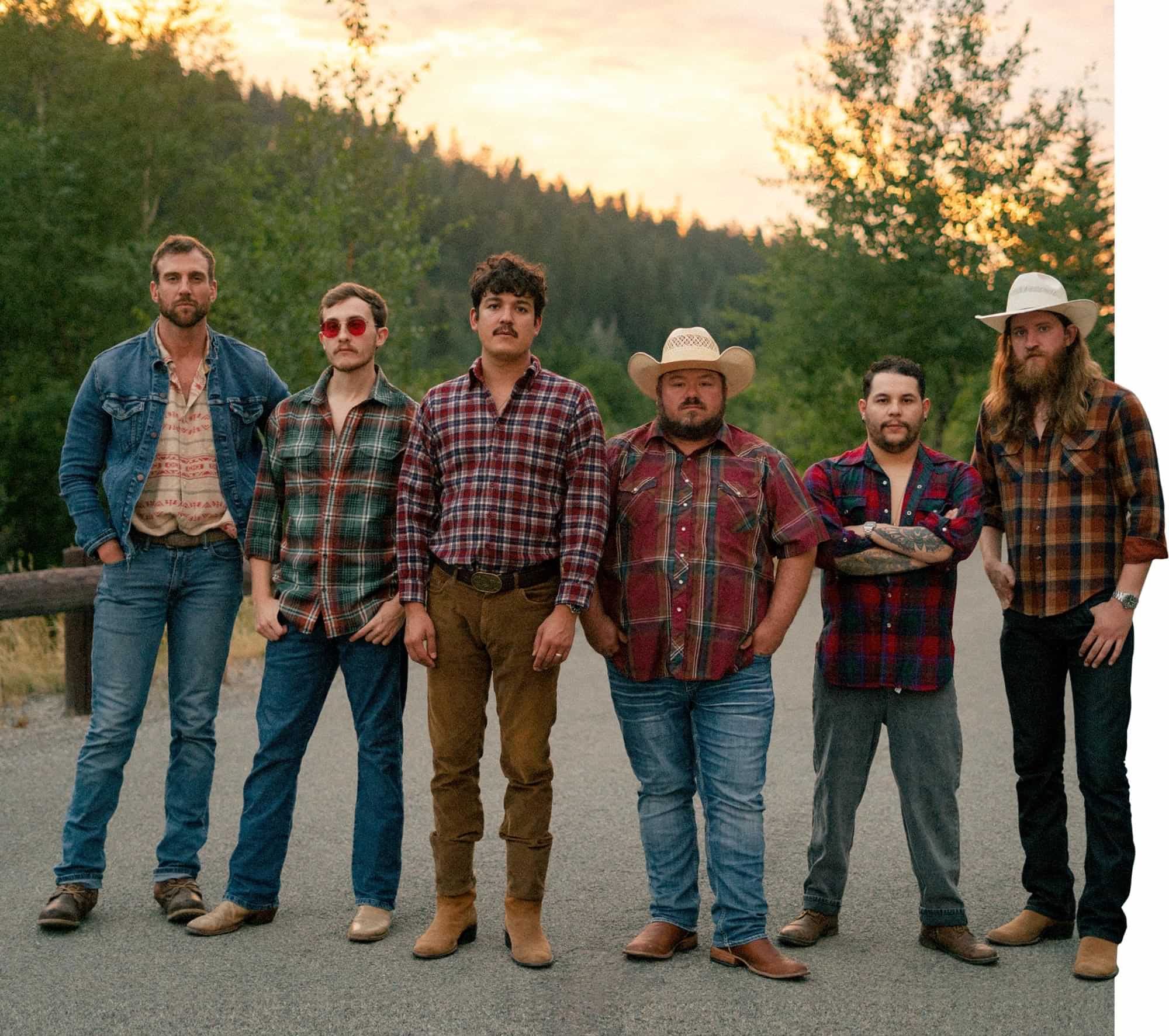 Jason Albers (far left) picture with his five bandmates, each wearing a different colored flannel shirt as they stand shoulder to shoulder on a road against the sun setting behind a high hill in the background