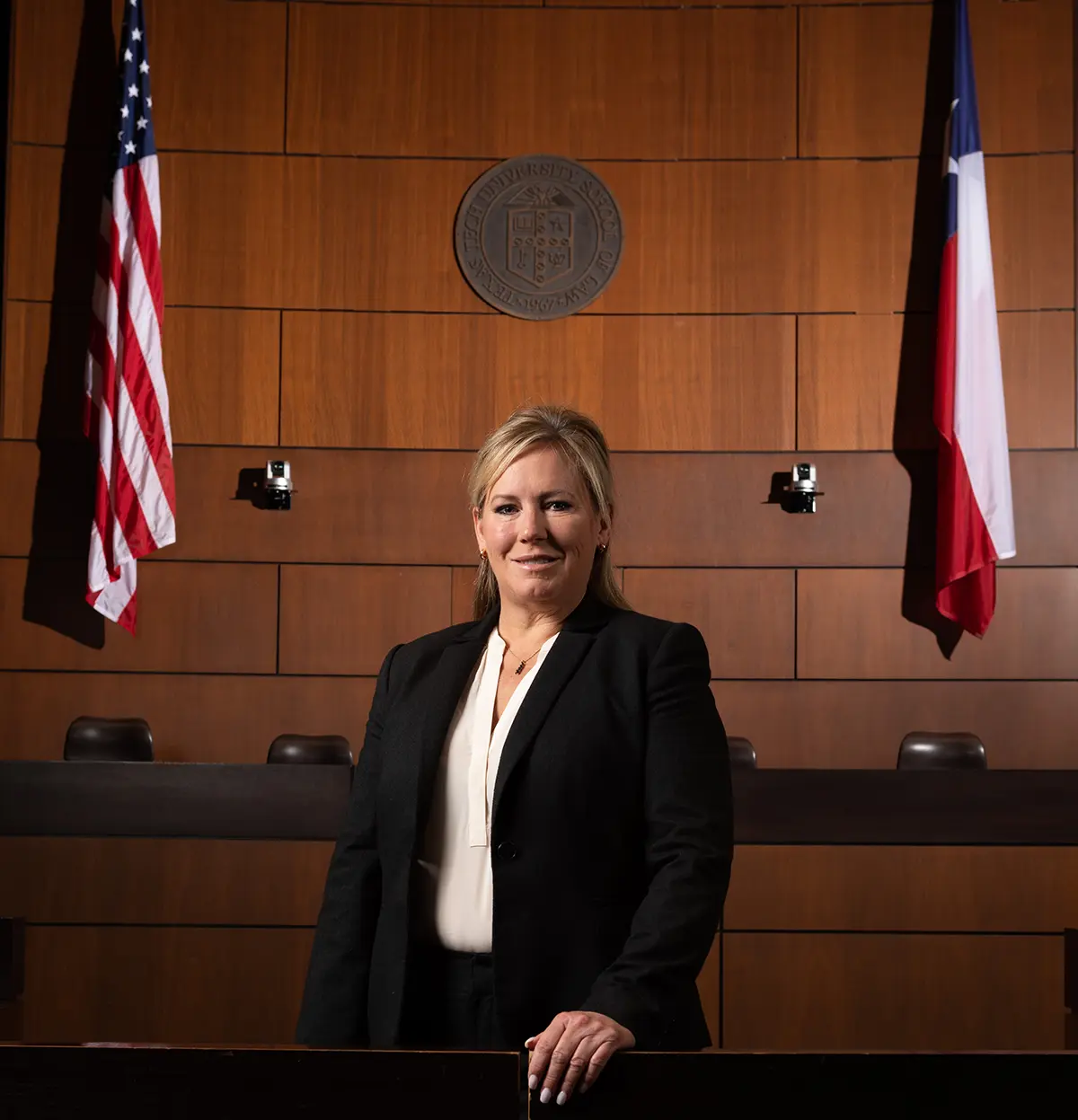 Portrait photograph of Carrie Edwards, PhD, CA- and CP-SANE, (Nursing ’93) smiling in a black open dress coat and white blouse underneath as she is inside a courtroom area at the Texas Tech University School of Law