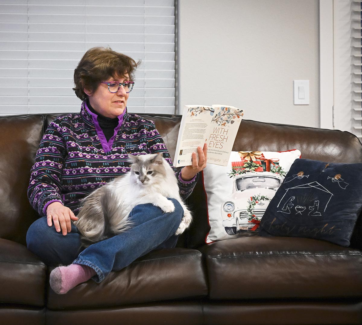 Susan Bergeson sitting cross-legged on the couch with a cat on her lap and reading a book