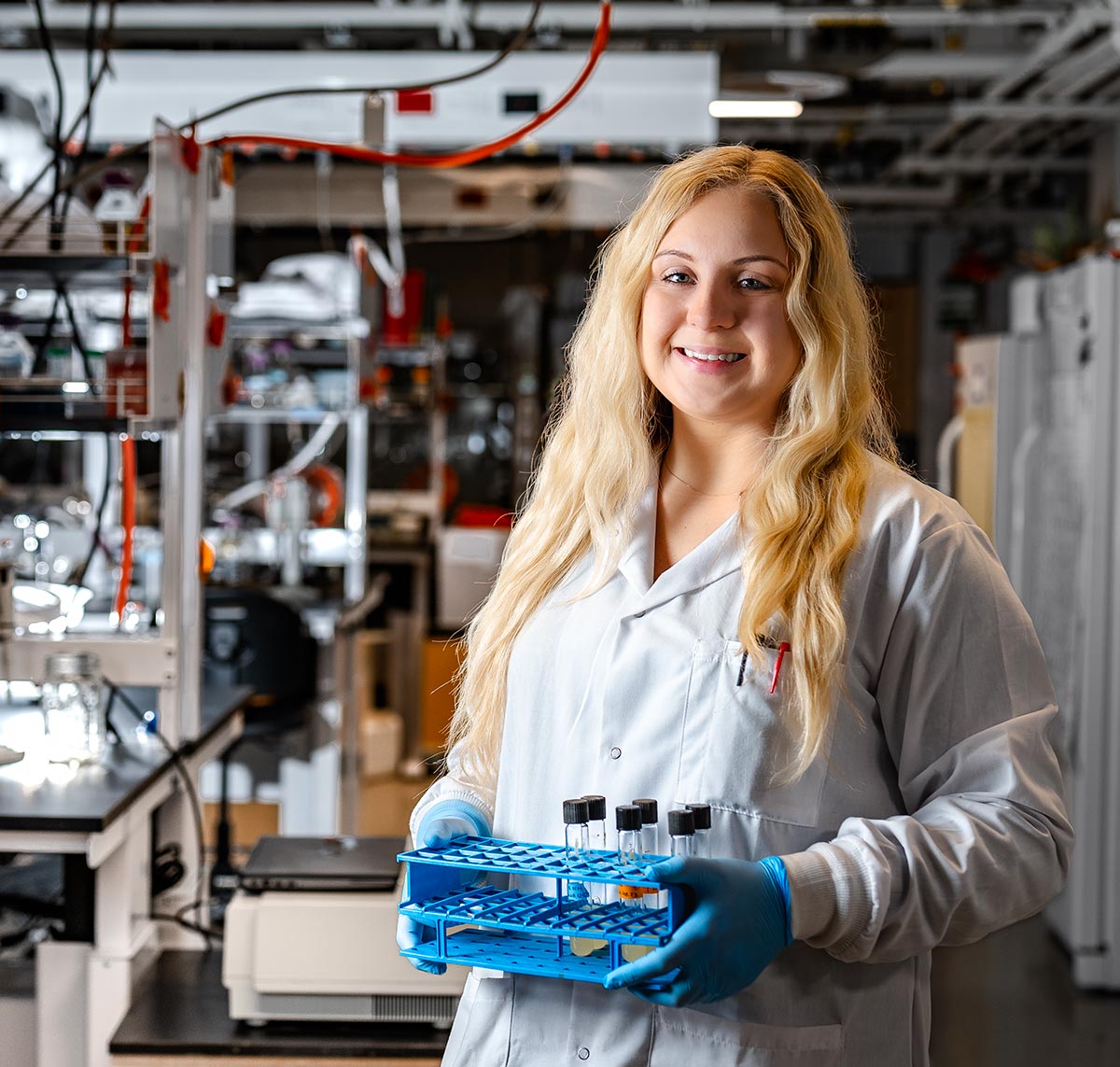 Portrait photograph of Whitni Redman (PhD, Biomedical Sciences '21 / Research Assistant Professor for Binghamton University's First-Year Research Immersion Program) smiling in a white lab coat holding a blue rectangular hole container with scientific laboratory collection tubes inside the container as she is standing somewhere inside a lab environment