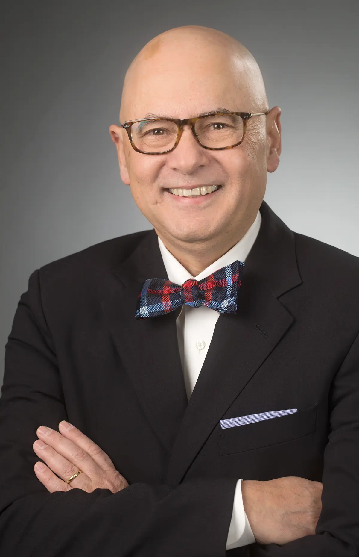 Portrait close-up headshot photograph of John C. DeToledo (MD) smiling in a black business suit blazer and a white button-up dress shirt underneath with a multi-colored (red/blue/white) pattern bowtie equipped as he has his arms crossed over each other