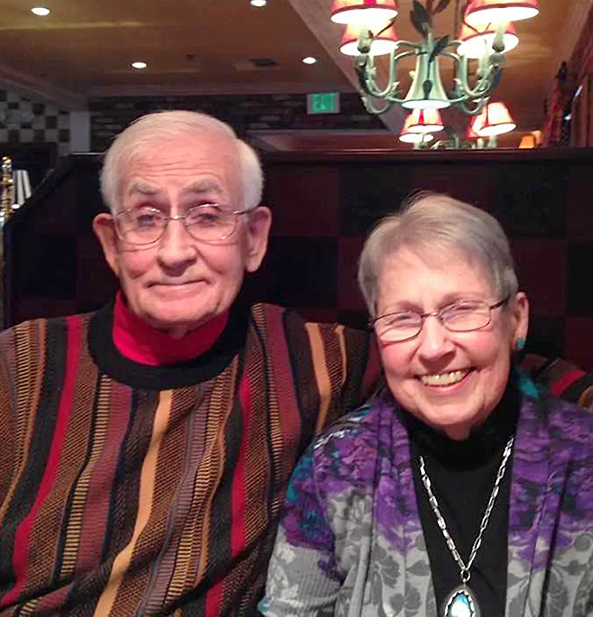 Portrait photograph close-up perspective of Darlene Norton in a open dark violet/grey colored cardigan jacket while she has on a black blouse underneath the cardigan and has on a chrome-colored necklace around her while her husband Bill Norton sits right next to her in a dark beige/black/red colored striped sweatshirt and chrome-colored outer frame prescription see through glasses as they are seated in what appears to be inside at a booth table within a restaurant setting