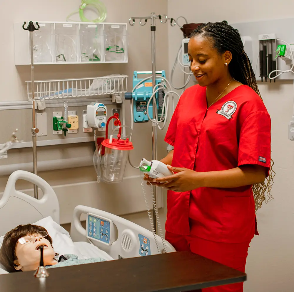 a female student dressed in vibrant red TTUHSC branded scrubs stands beside a patient care training simulator laying in a hospital bed