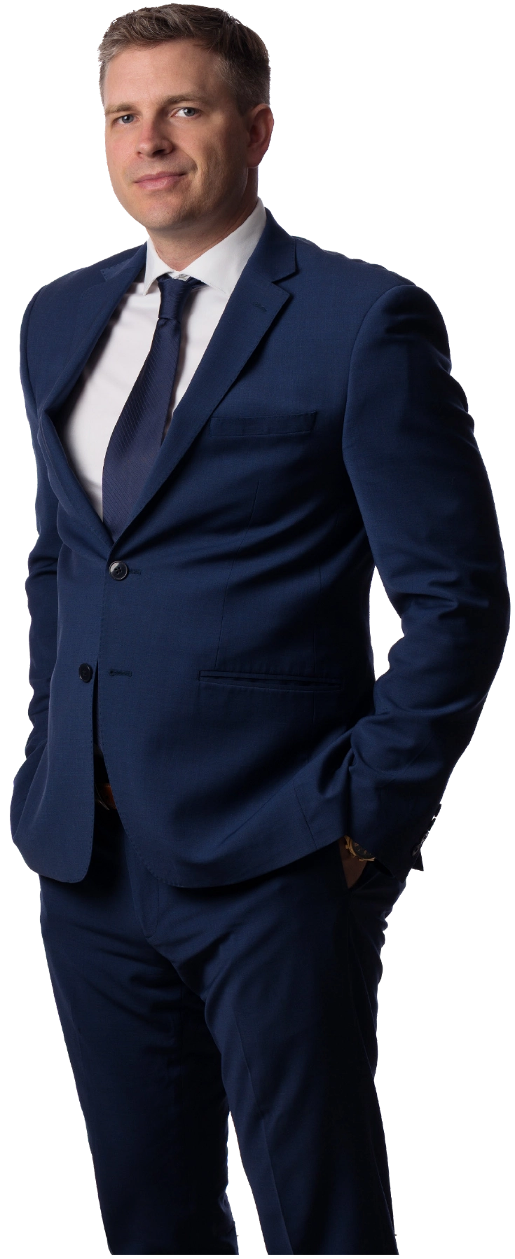 A mid-body portrait photograph of Aric Logsdon, PhD, a man with short dark brown hair, wearing a navy blue business blazer suit outfit, white button-up dress shirt, and navy blue tie, standing with his hands in his pockets as he has a slight faint grin