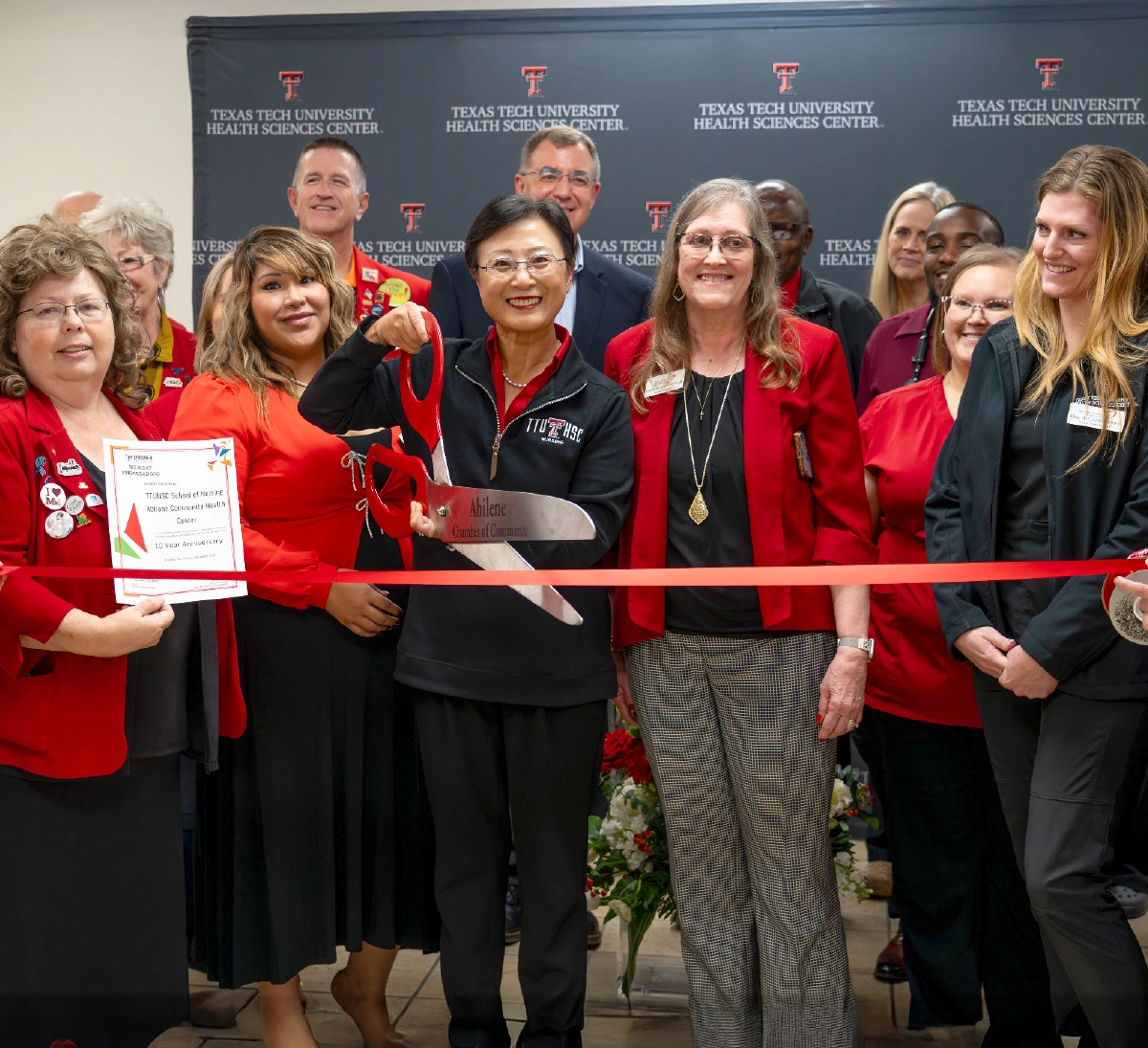 A large group of Texas Tech University Health Sciences Center staff and faculty individuals gathered for a celebration; In the center, a woman in a black and red jacket holds oversized red scissors to cut a red ribbon; A woman to the left holds a framed certificate that reads 10 Year Anniversary; The background features a banner for Texas Tech University Health Sciences Center