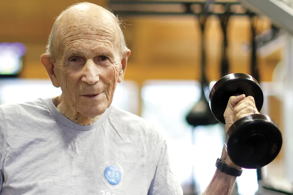 Elderly man exercising in a gym while lifting a dumbbell, wearing a gray T-shirt with a Cooper Clinic fitness emblem and the year 2025, demonstrating strength training and active aging.