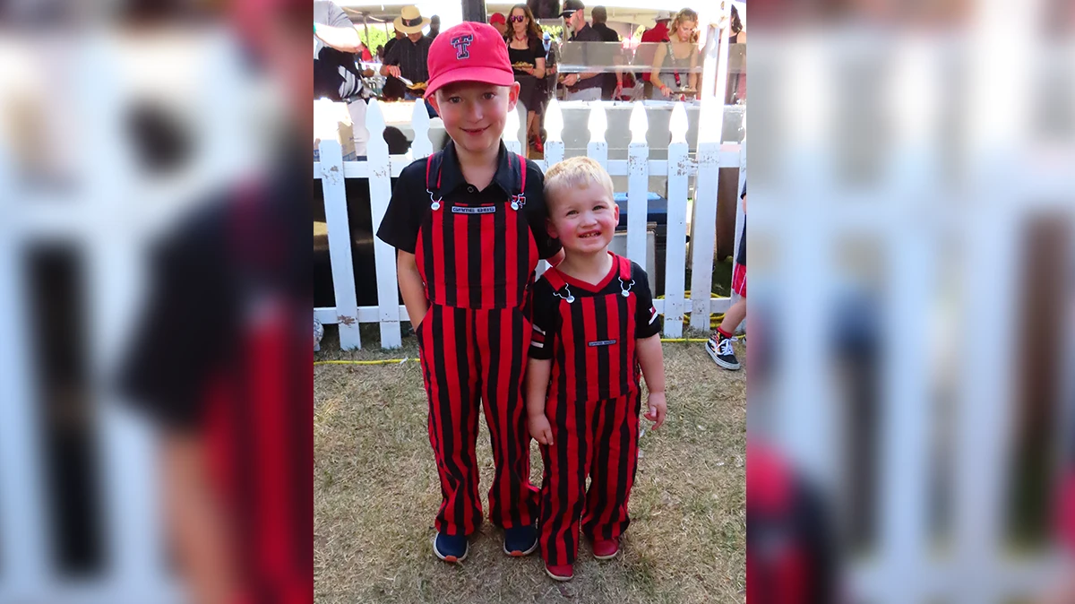 Two children wearing matching red-and-black striped overalls stand side by side at an outdoor event, smiling in front of a white picket fence with a crowd in the background.