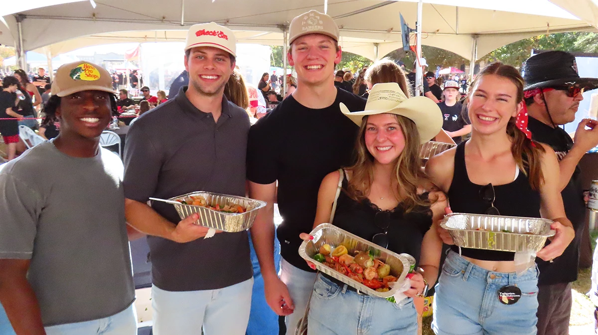 Group of young adults smiling at an outdoor tailgate-style event while holding aluminum food trays, standing under a large tent with other attendees behind them.