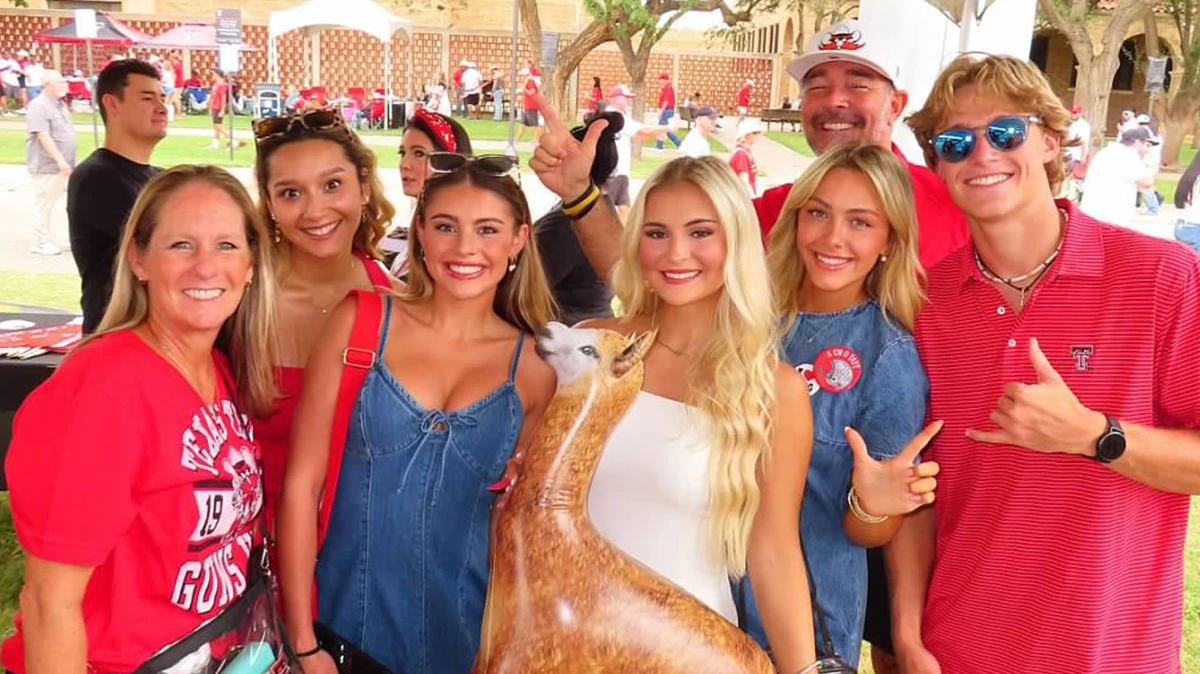 Group of people posing together at an outdoor event, smiling and wearing red and black clothing, with a large decorative longhorn statue positioned in front of them.