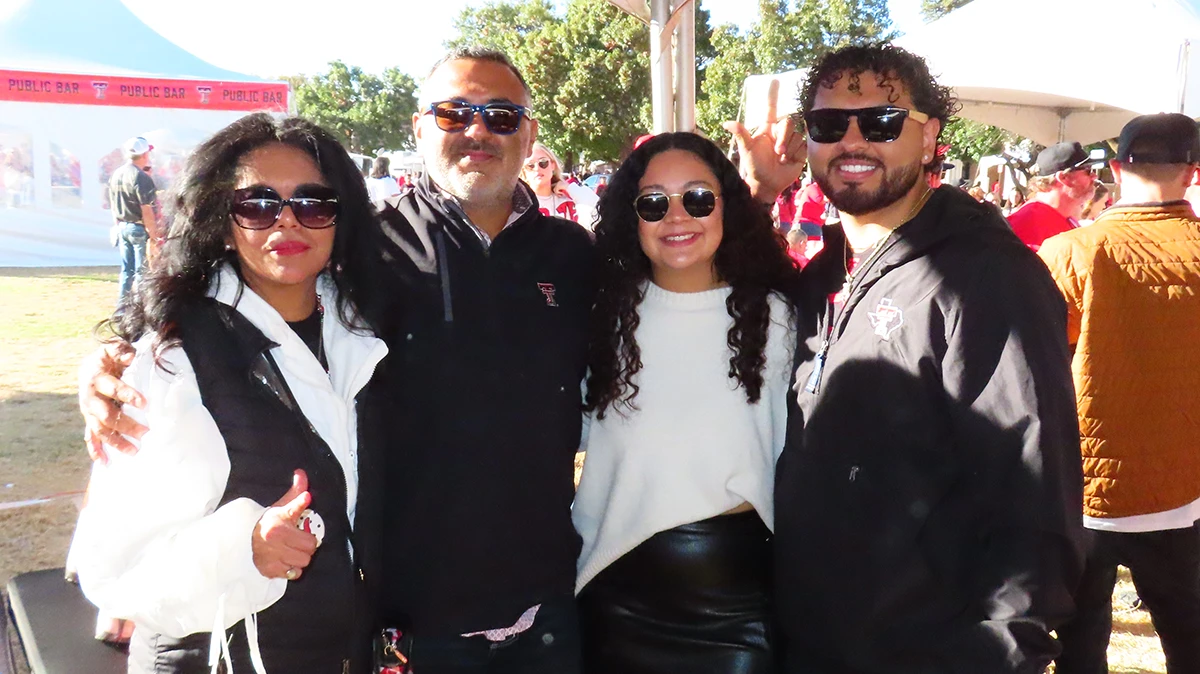 Four people posing closely together at an outdoor gathering, smiling and wearing casual jackets and sunglasses under a large event tent.