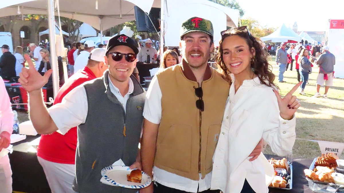 Three people standing together at an outdoor event, smiling and holding plates of food, with tents, tables, and other attendees visible behind them.