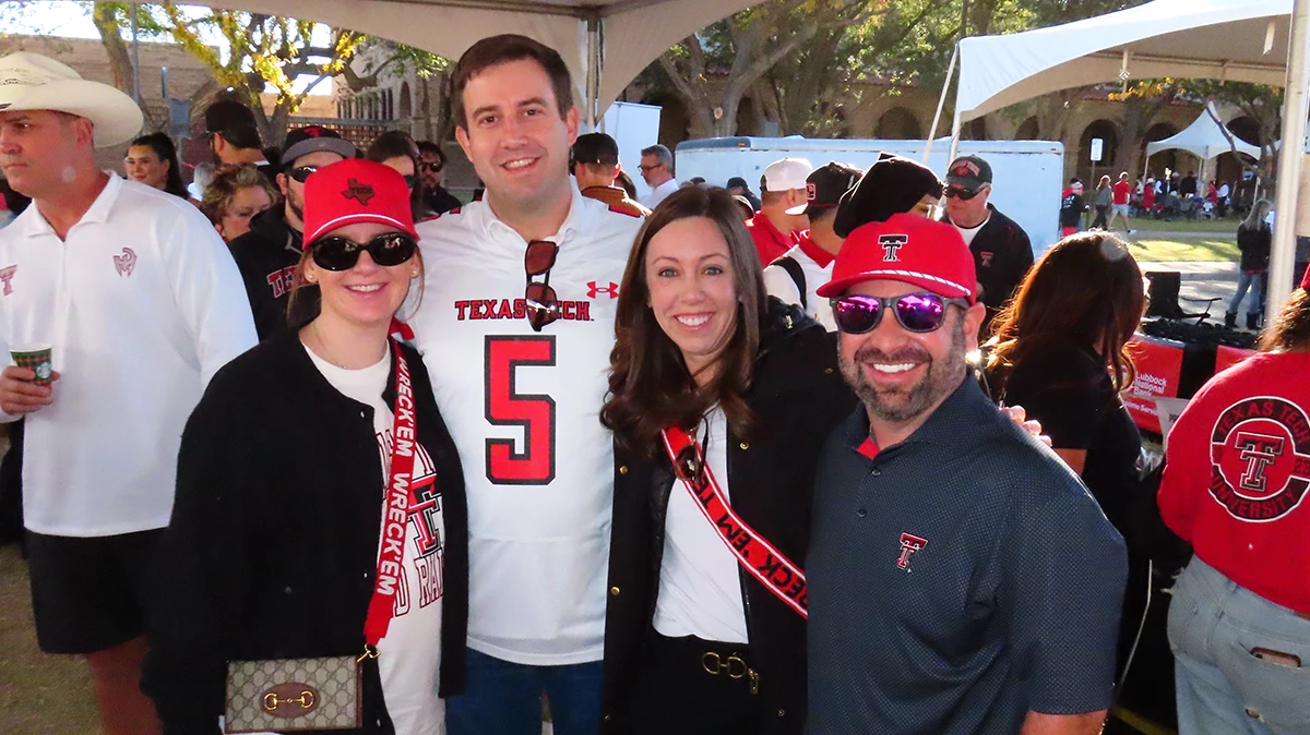 Group of five people posing together at an outdoor event, including one person seated in a wheelchair, smiling and holding drinks near a tented food area.
