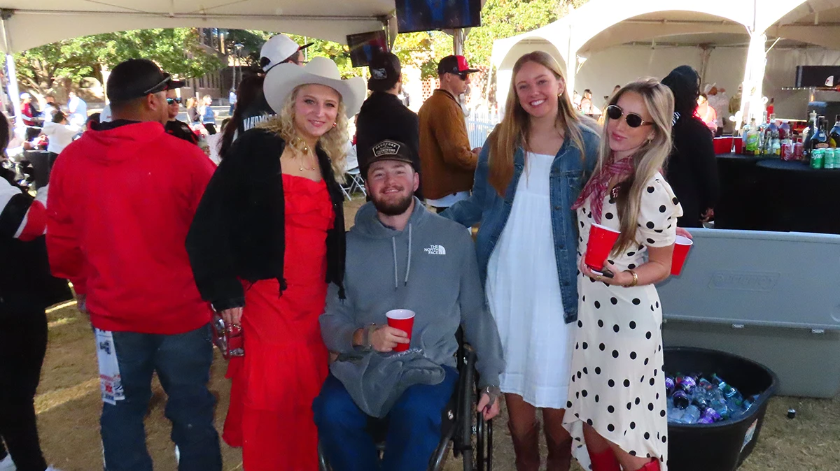 Four people standing together at an outdoor event, smiling and wearing casual clothing, with food tables and tents visible in the background.