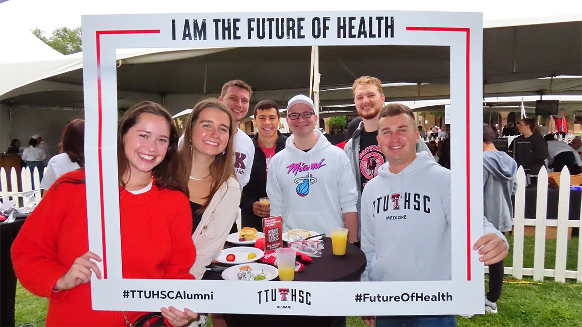 Group of young adults posing behind a large photo frame that reads “I Am the Future of Health,” smiling at an outdoor alumni event with tents and tables nearby.
