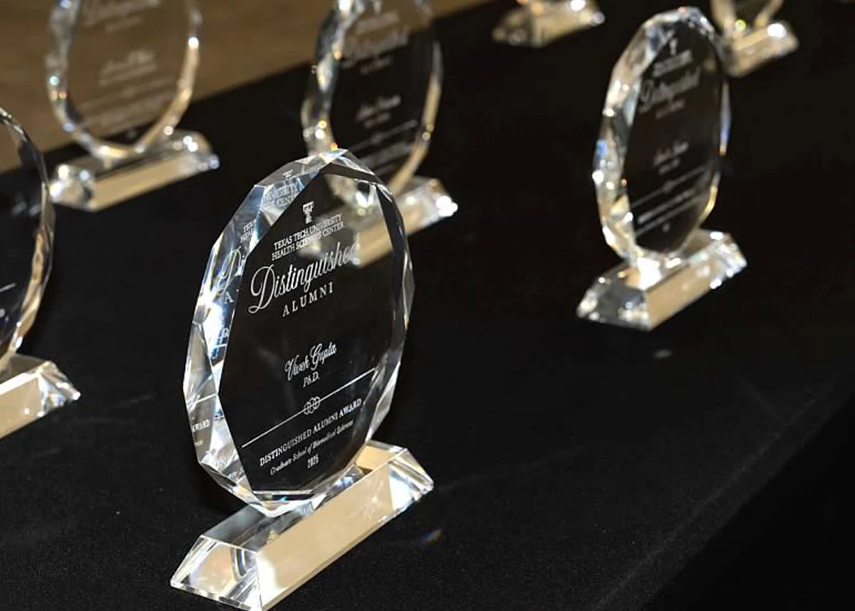 Close-up view of multiple clear crystal awards arranged on a black tablecloth, engraved with “Distinguished Alumni” and Texas Tech University Health Sciences Center branding.
