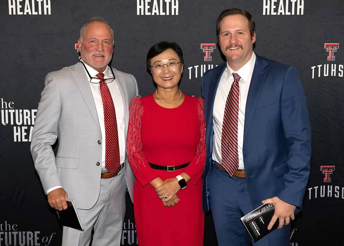 Three adults dressed in formal attire posing together in front of a “The Future of Health” TTUHSC backdrop, with one person holding a Distinguished Alumni award.