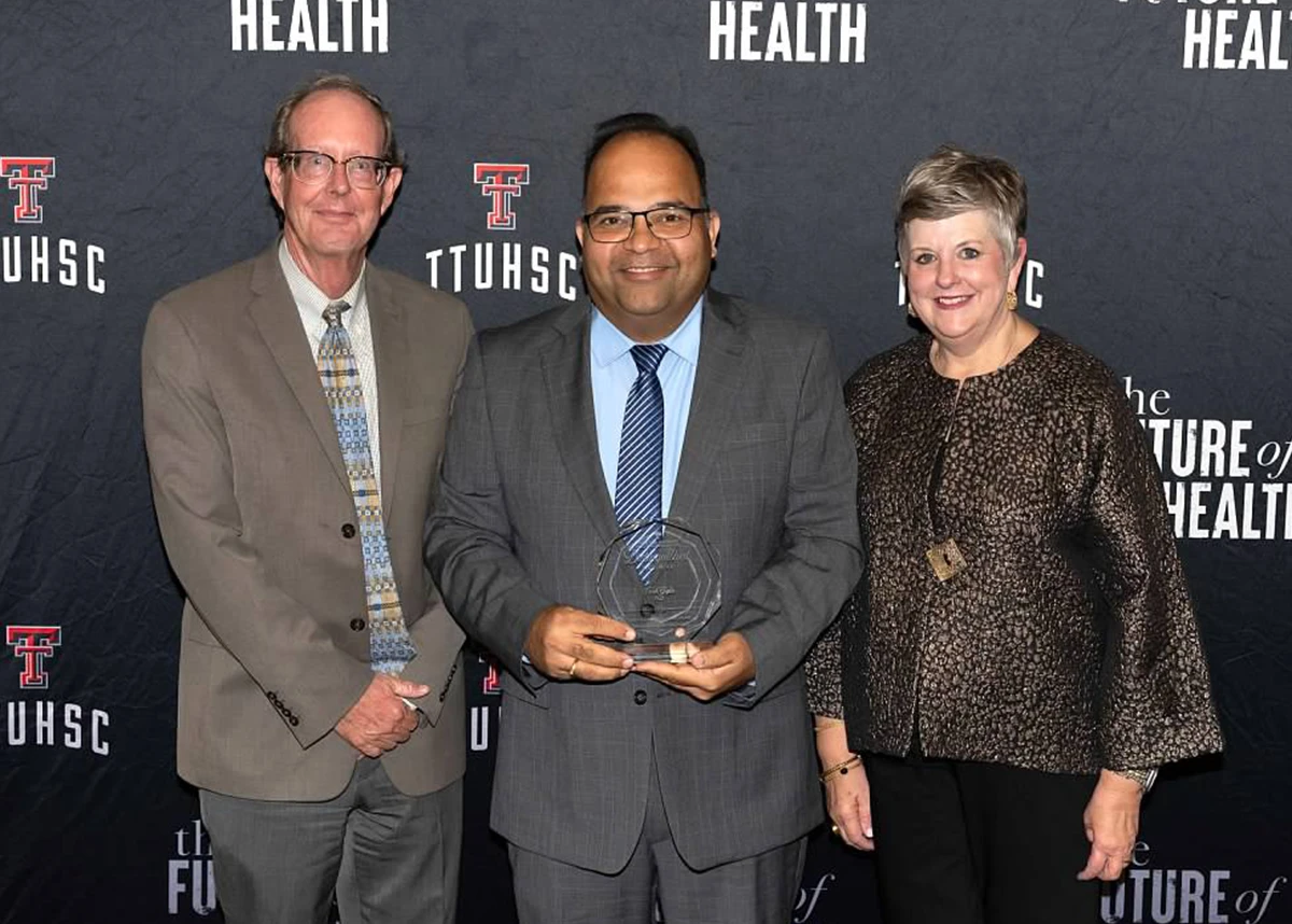 Three adults dressed in formal attire pose in front of a TTUHSC “The Future of Health” backdrop, holding a Distinguished Alumni award at an event.