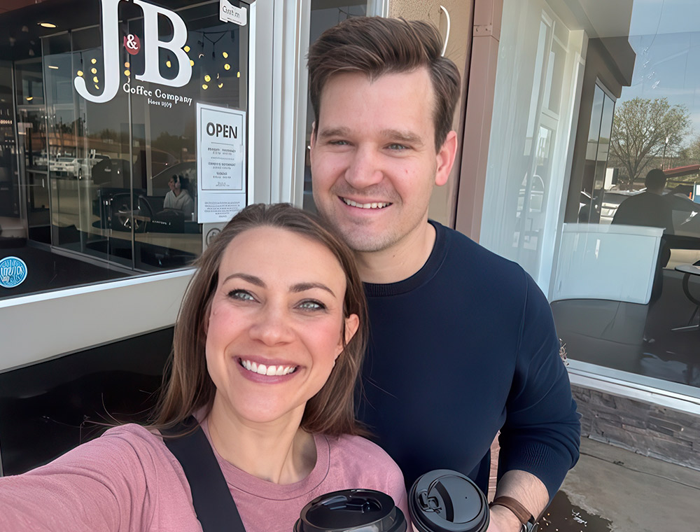 DJ and Tyler Sutton smile for a selfie outside J&B Coffee Company, each holding a coffee cup. The coffee shop's logo and an "OPEN" sign are visible in the background on the glass storefront.