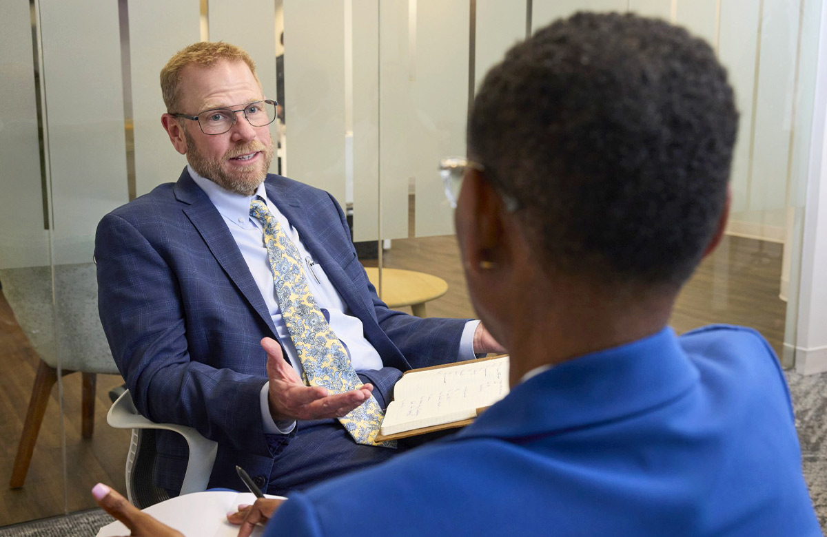 Bob Dent, wearing a suit and glasses, gestures with his hand while speaking to a person in a blue blazer in an office with glass walls. He has an open notebook on his lap.