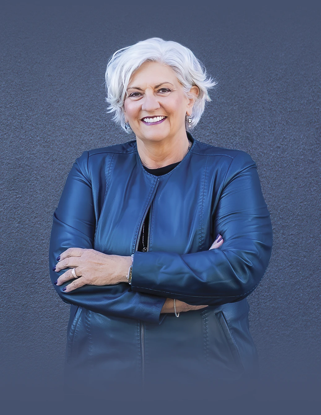 Portrait of a smiling woman with short white hair standing against a dark gray wall, wearing a blue jacket with her arms crossed, conveying a confident and professional demeanor.