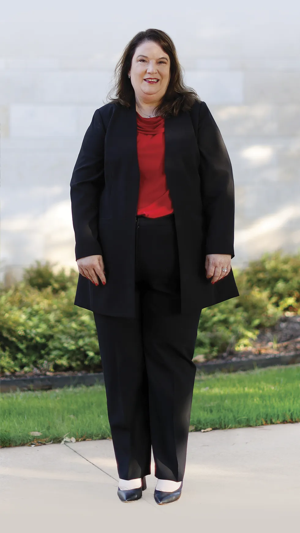 Full-length portrait of a woman standing outdoors on a paved walkway, wearing a black suit with a red top, posed in front of a light stone wall and landscaped greenery.