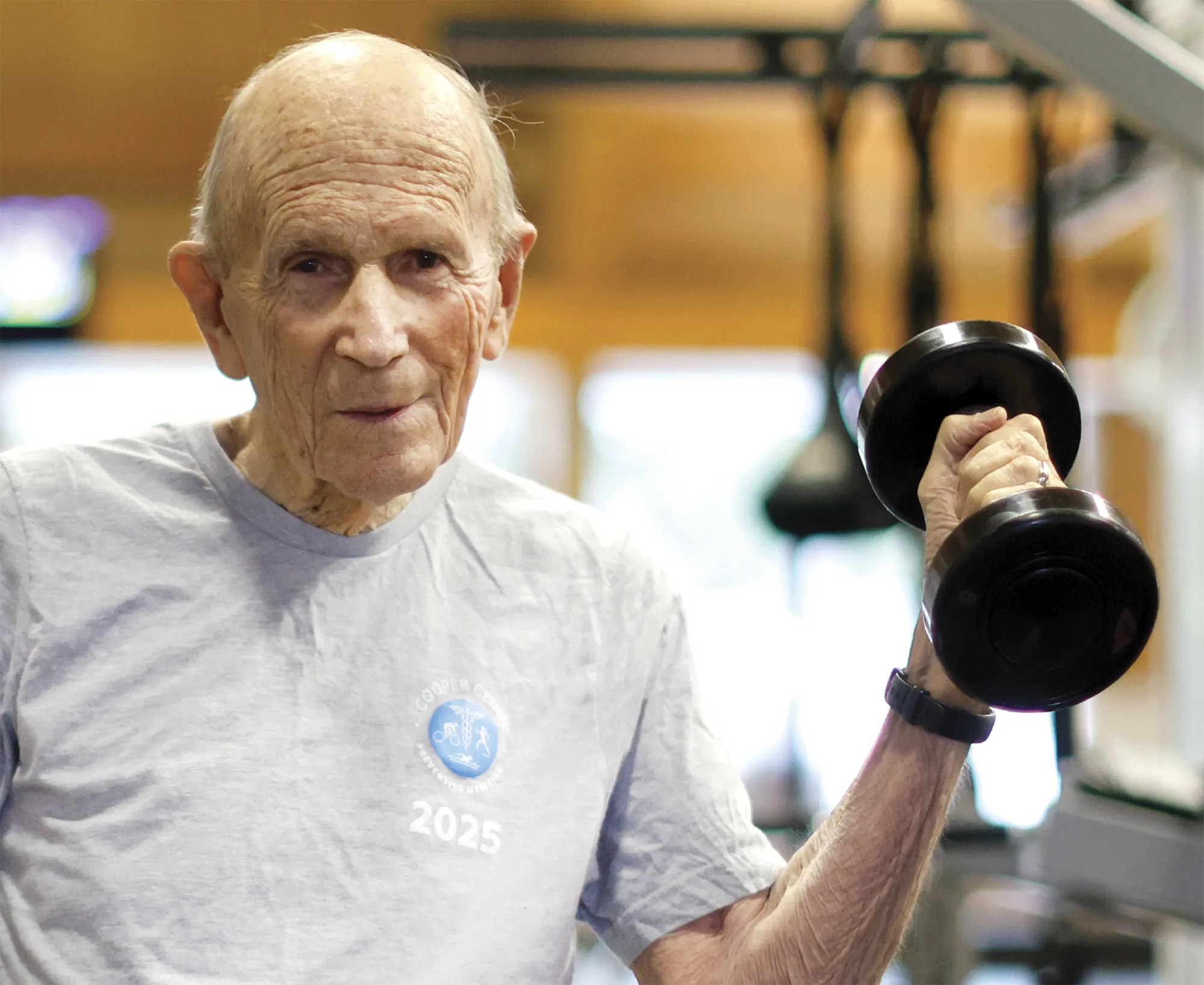 Elderly man exercising in a gym while lifting a dumbbell, wearing a gray T-shirt with a Cooper Clinic fitness emblem and the year 2025, demonstrating strength training and active aging.