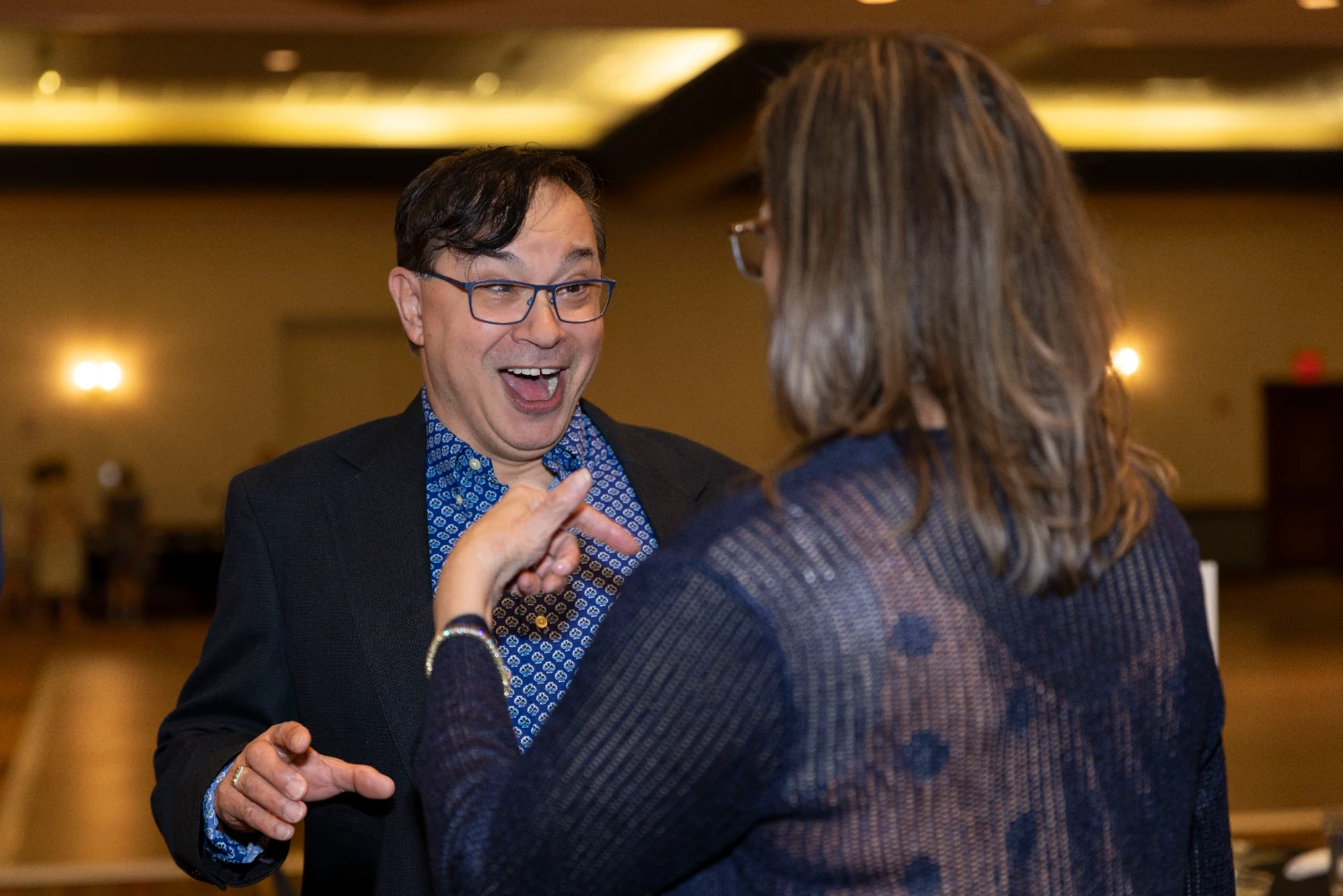 A man reacts enthusiastically while speaking with a woman at an indoor event.