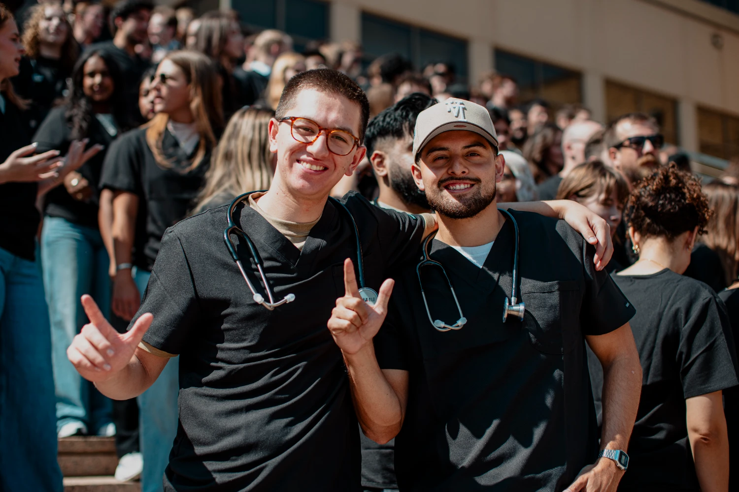 Two medical students wearing scrubs and stethoscopes pose together and make the Guns Up hand sign.