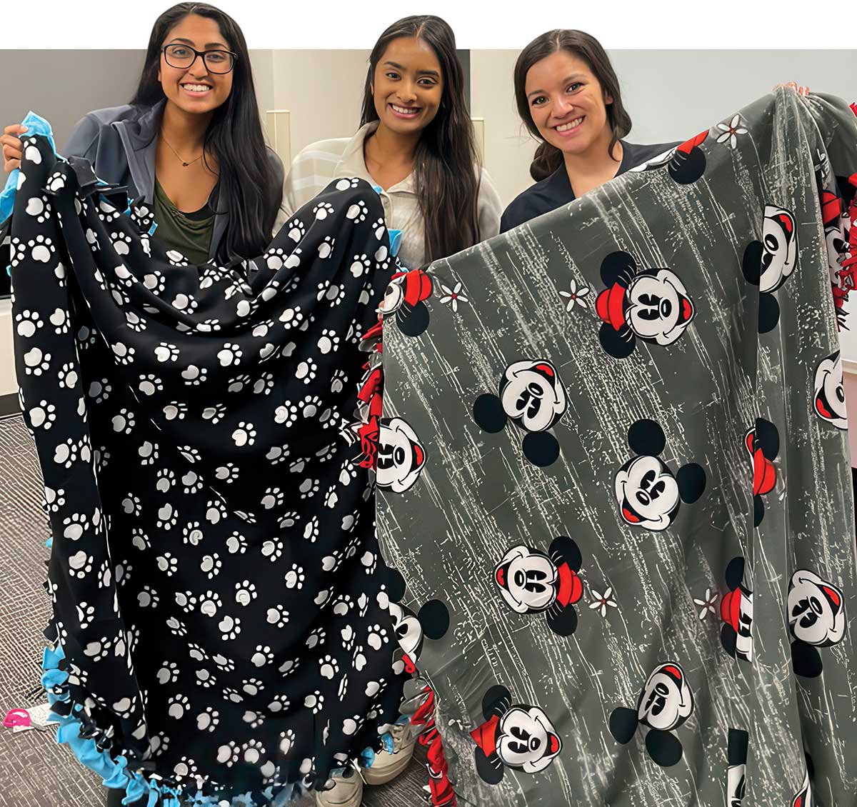 Three smiling women display two patterned fleece blankets, one with paw prints and one with Mickey Mouse.