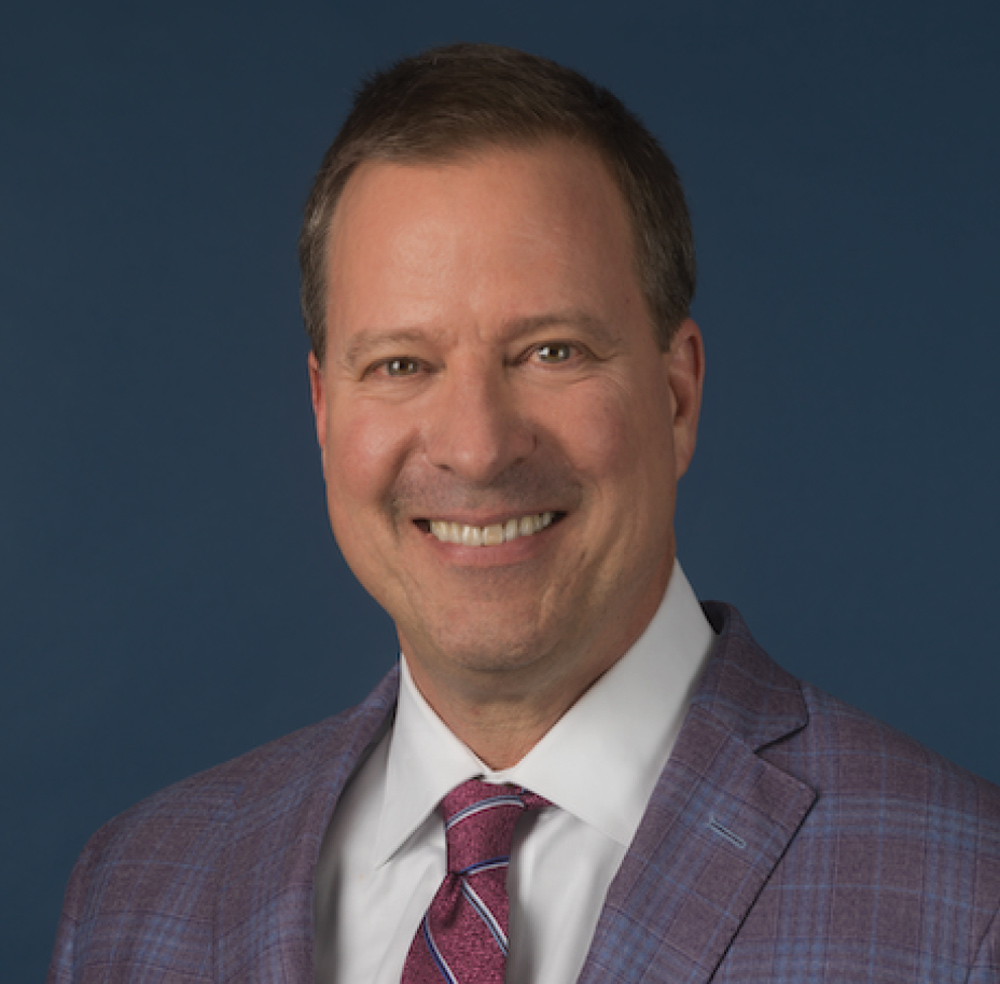 Headshot of a smiling Lance R. McMahon with short brown hair, wearing a patterned blue blazer, white shirt, and purple tie.