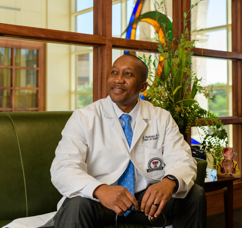 Izi Obokhare wearing a white lab coat and blue tie sitting on a green couch in a modern office.