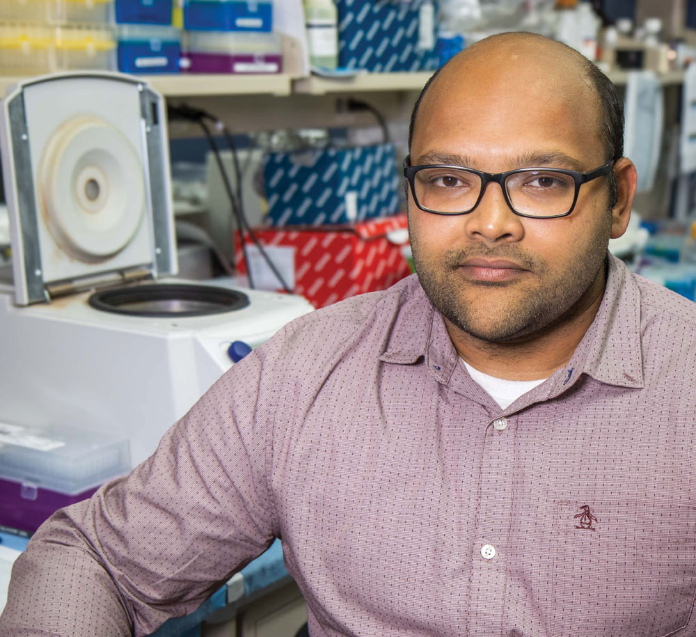 Balakrishna Koneru wearing glasses and a button-down shirt sitting in a laboratory next to an open centrifuge machine.
