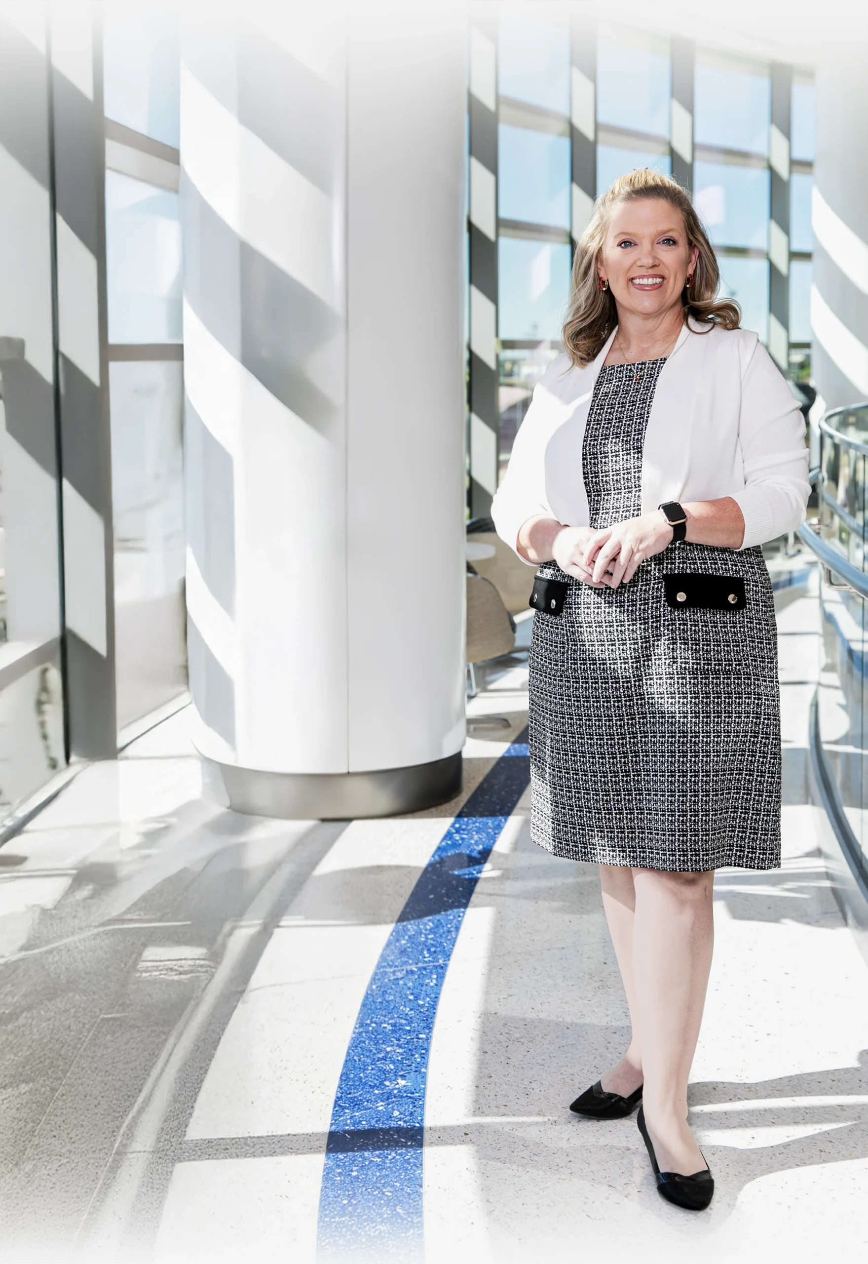 A woman in professional attire stands in a sunlit corridor with large windows and a curved blue floor detail.