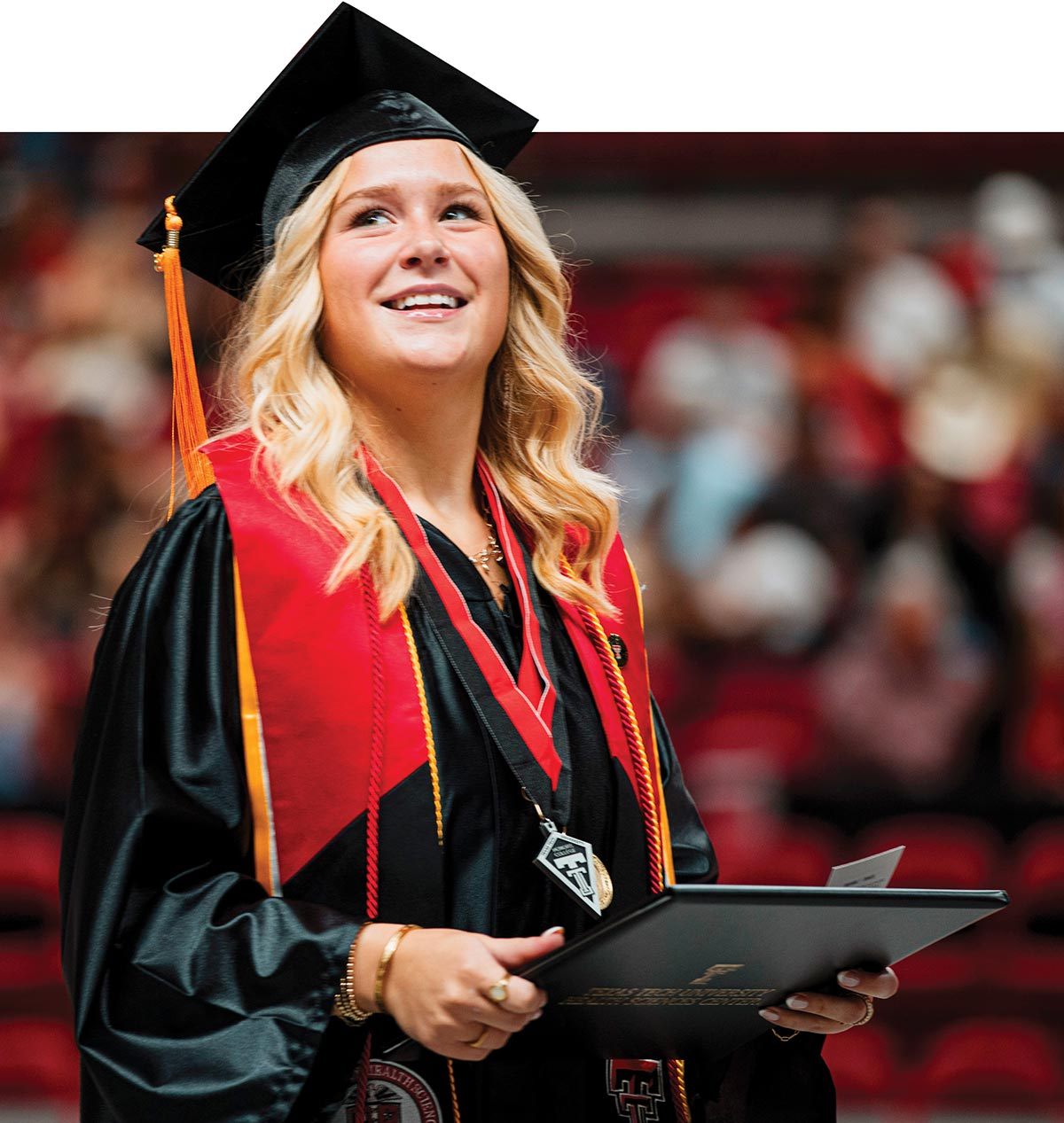 A smiling Texas Tech graduate wearing a cap, gown, and red stole, looking upward while holding her diploma cover during a commencement ceremony.