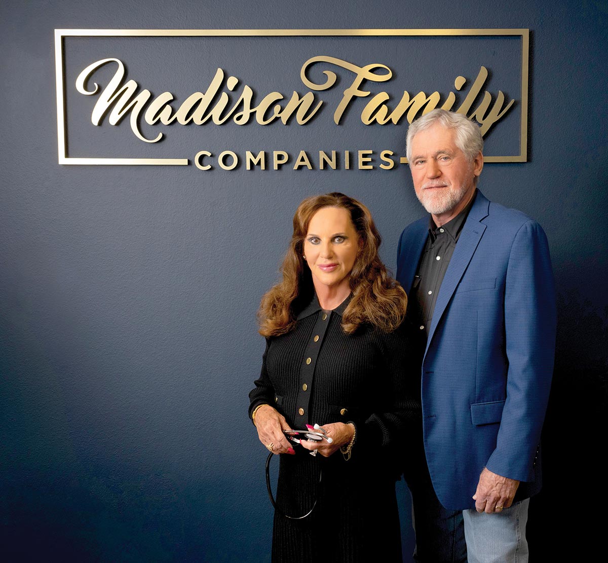 A man and woman posing together in front of a gold "Madison Family Companies" sign on a dark blue wall, with the woman holding a stethoscope.