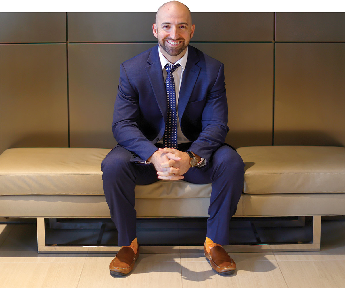 A smiling bald man wearing a blue suit, polka-dot tie, and bright yellow socks, sitting on a beige leather bench with hands clasped.